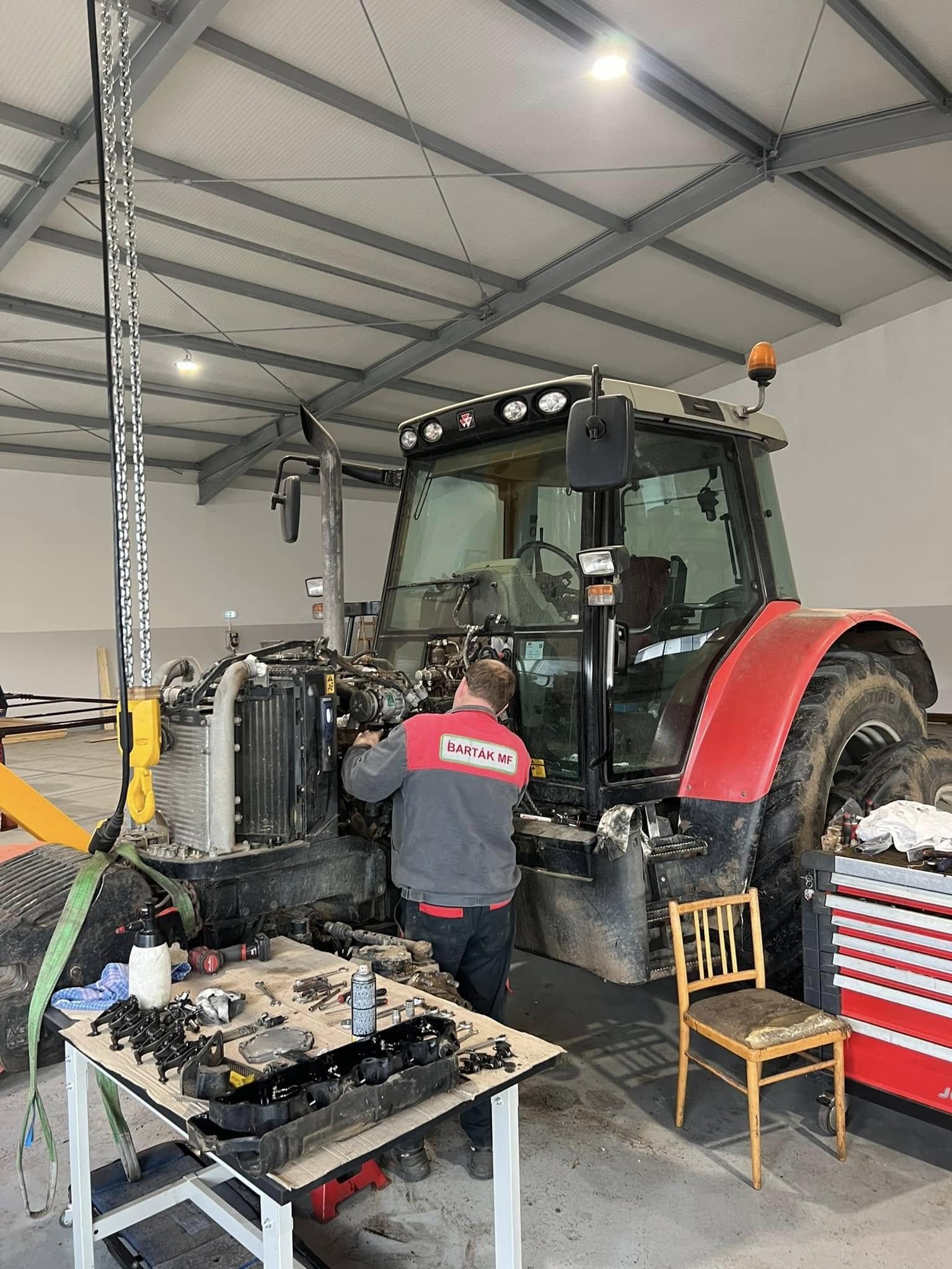 Mechanik opravuje traktor v dílně, vedle pracovní stůl s rozebranými díly.